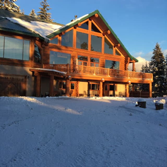 A wooden lodge covered in snow with large windows and a snowy landscape, located at Spirit Lodge in Vernon, BC, under a clear blue sky.