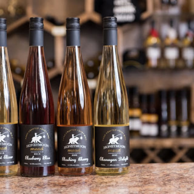Four bottles of Honeymoon Mead in different flavors are displayed on a countertop with a blurred background of shelves with other bottles.