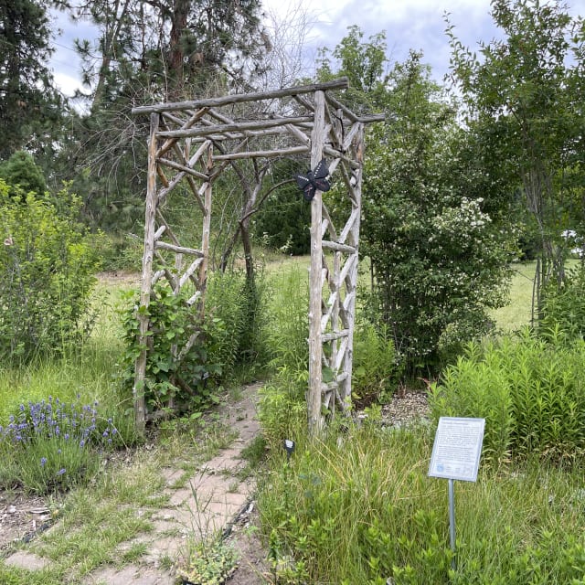 A rustic wooden garden archway with a butterfly decoration leads to a path surrounded by lush greenery and flowers in Summerland.