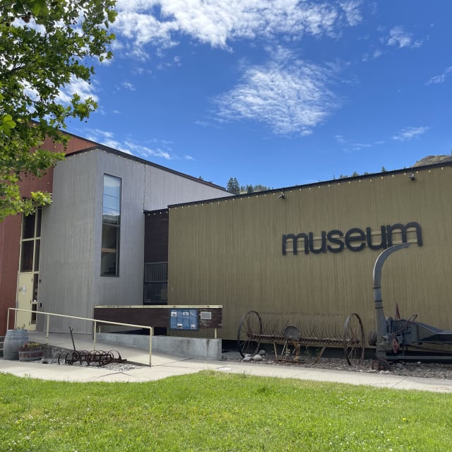 A modern building with a green facade and sign "museum" displayed outside, featuring rustic machinery, set against a clear blue sky in Summerland.