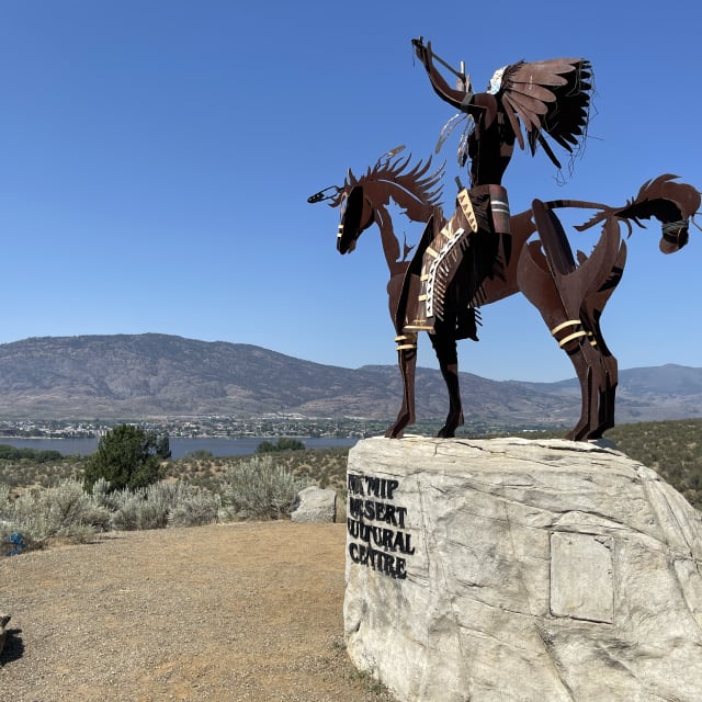 Metal sculpture of a Native American on horseback at Nk'Mip Desert Cultural Centre with Osoyoos and mountains in the background.