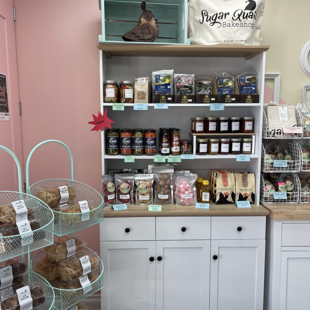 Shelf with various local products including jams, coffee, and pastries from Sugar Ruai Bakeshop in Osoyoos, with a teal metal stand displaying bakery items.