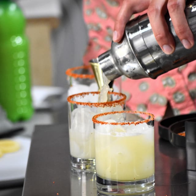 A person is pouring a beverage from a shaker into glasses rimmed with spice, set on a metal surface at Controlled Entropy Distilling in Summerland.