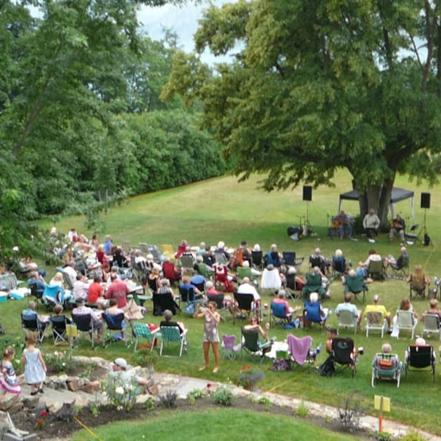 A crowd seated on lawn chairs at an outdoor musical event in Vernon, surrounded by trees on a sunny day.