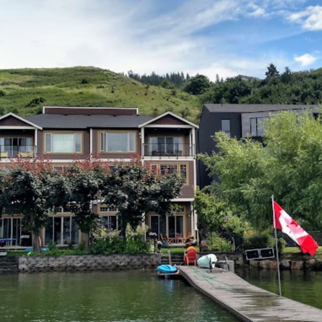 Charming lakeside house in Vernon surrounded by greenery, with a dock and Canadian flag in the foreground under a blue sky.