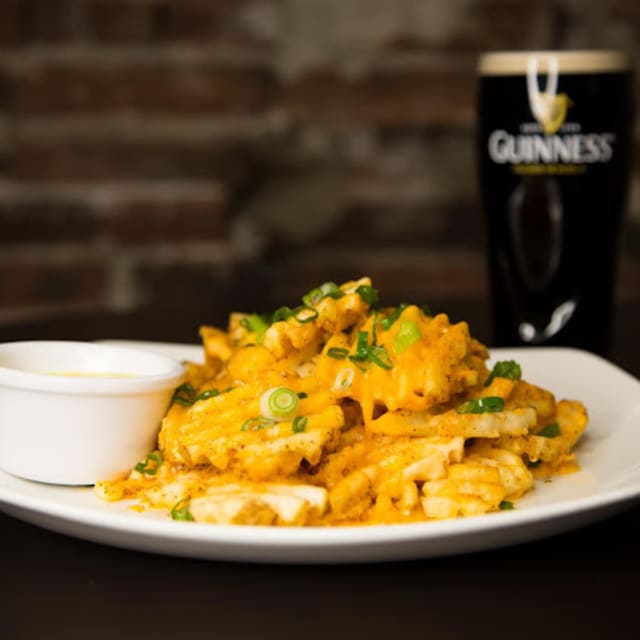 A plate of waffle fries topped with melted cheese and green onions, served with a dip, is placed in front of a pint of Guinness on a dark table.