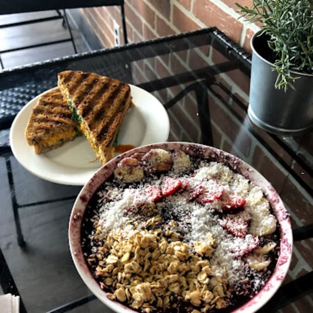 Açai bowl with granola, strawberries, bananas, and coconut beside a grilled sandwich on a glass table with a brick wall and plant in Vernon.