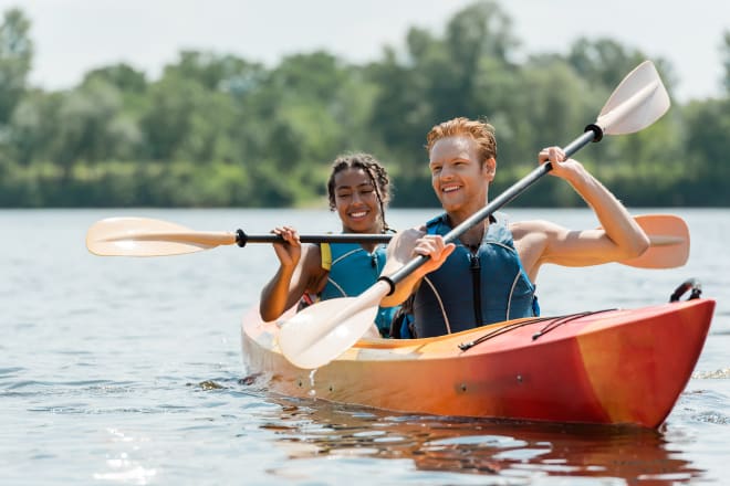 Two people kayaking on a lake with green trees in the background.