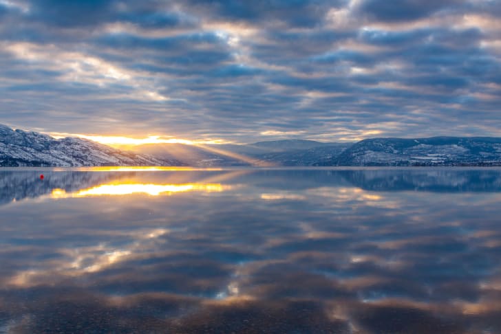 A serene sunset over a calm lake with surrounding snow-capped mountains under a cloudy sky in Kelowna.