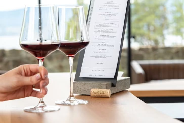 Two glasses of red wine held by a hand with a CedarCreek Estate Winery wine menu on a table.