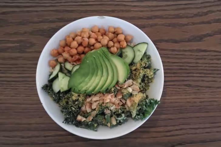 A bowl of colorful salad with avocado slices, chickpeas, cucumber, and ingredients on a wooden table.