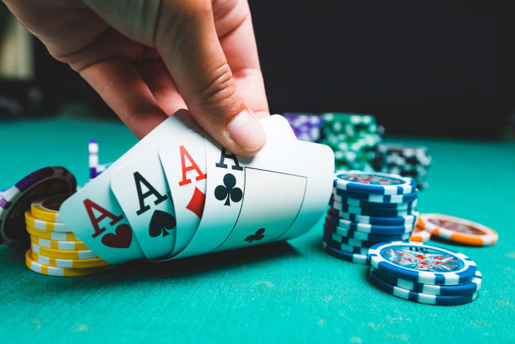 A hand holding four aces over a green casino table with poker chips in the background.