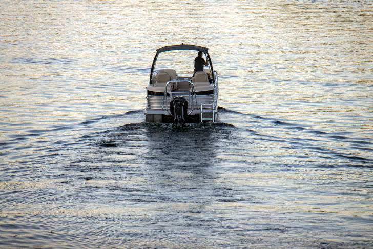 A person driving a pontoon boat on calm lake waters during a sunny day.