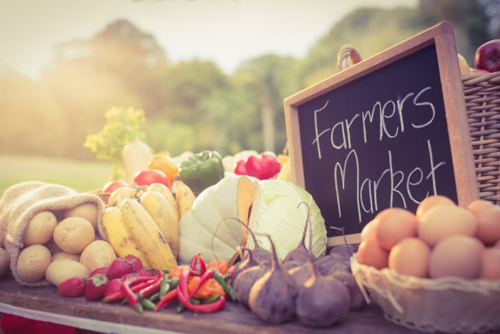 Fresh produce including potatoes, bananas, peppers, cabbage, eggs, and more displayed at a Farmers Market with a chalkboard sign.