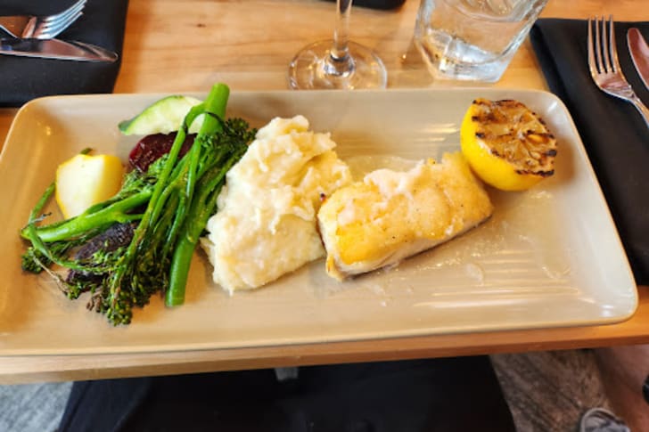 A rectangular plate with a serving of mashed potatoes, grilled fish fillet, steamed broccoli, a charred lemon half, and other side vegetables, with a glass of water and cutlery beside it.