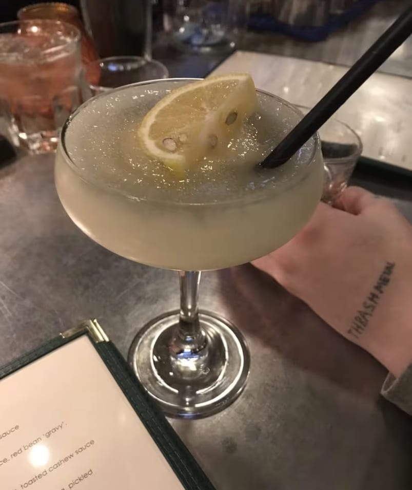 A frosty cocktail with a lemon garnish and straw on a table at BNA Brewing in Kelowna, with a hand holding the glass and a menu partially visible.