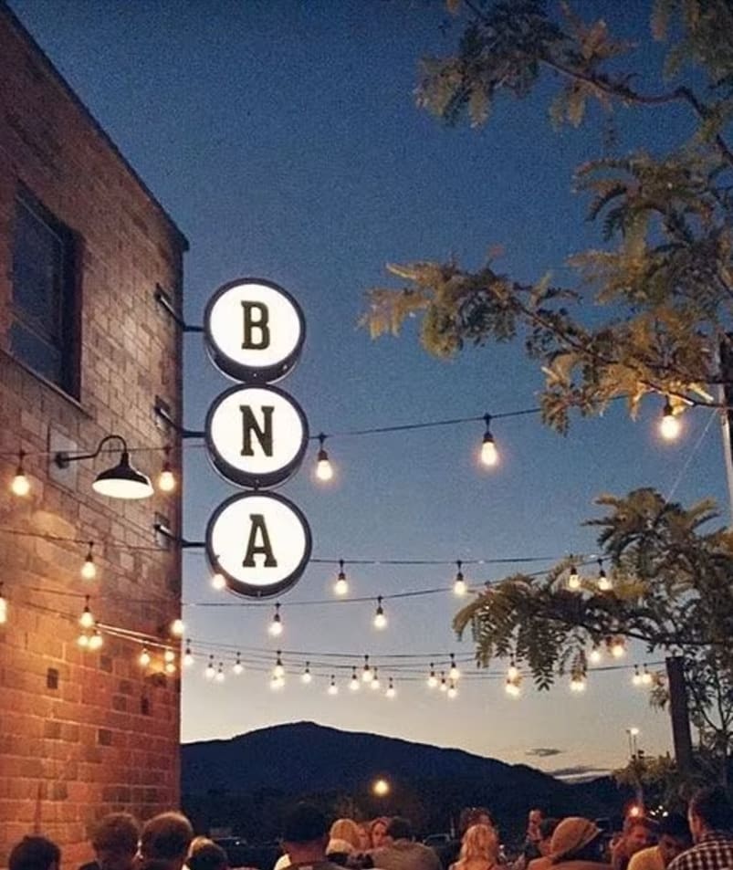 A gathering of people at BNA Brewing in Kelowna during the evening with string lights hanging above and a BNA sign illuminated on the brick building.