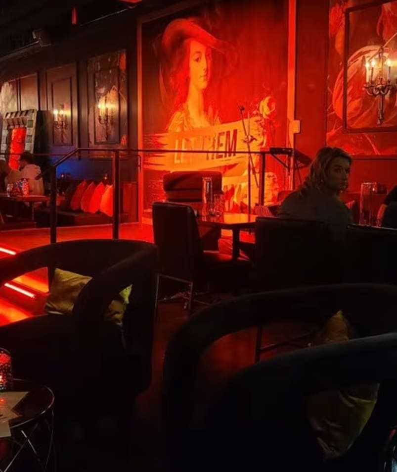 A group of people sitting at tables in a room with red and blue lights on the walls and a painting on the wall at Friends of Dorothy Lounge in Kelowna.
