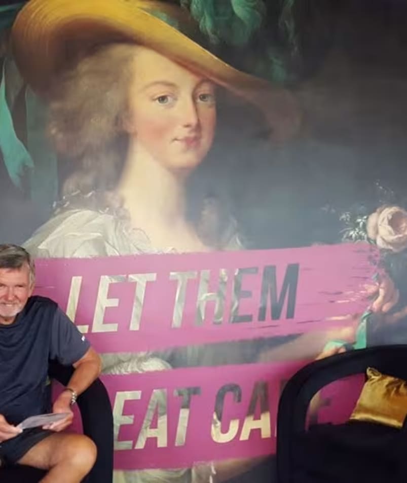 A man sitting on a chair in front of a painting of a woman with a hat on her head at Friends of Dorothy Lounge in Kelowna.