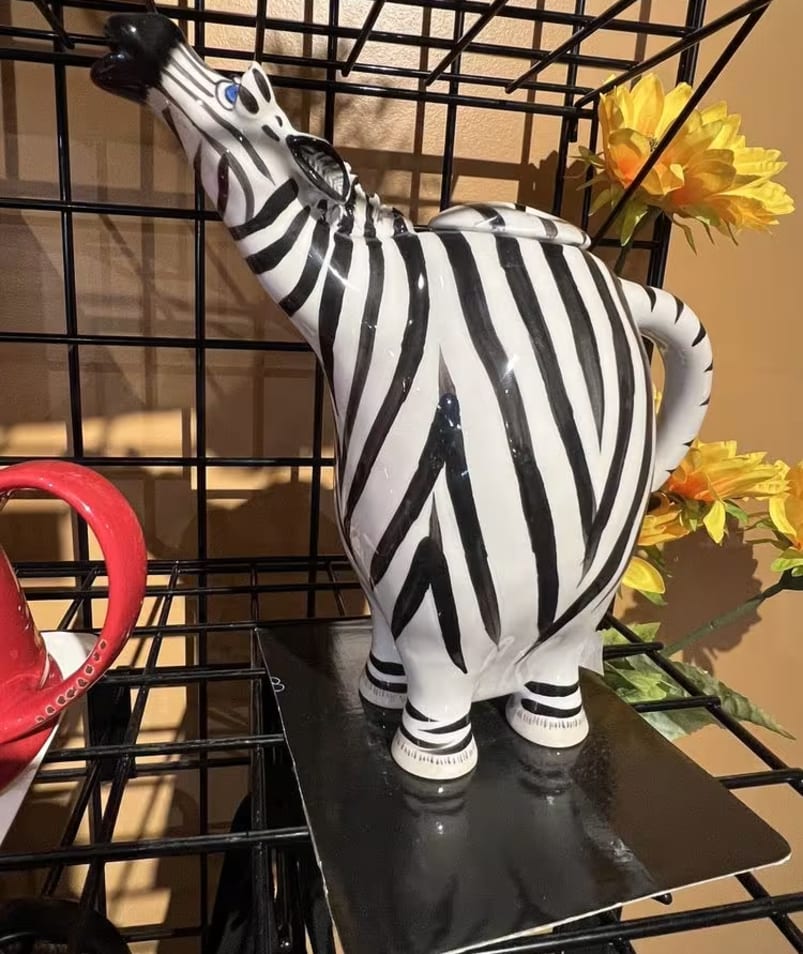 A zebra-shaped teapot displayed on a black metal shelf with yellow flowers in the background at The Jammery in Lake Country.