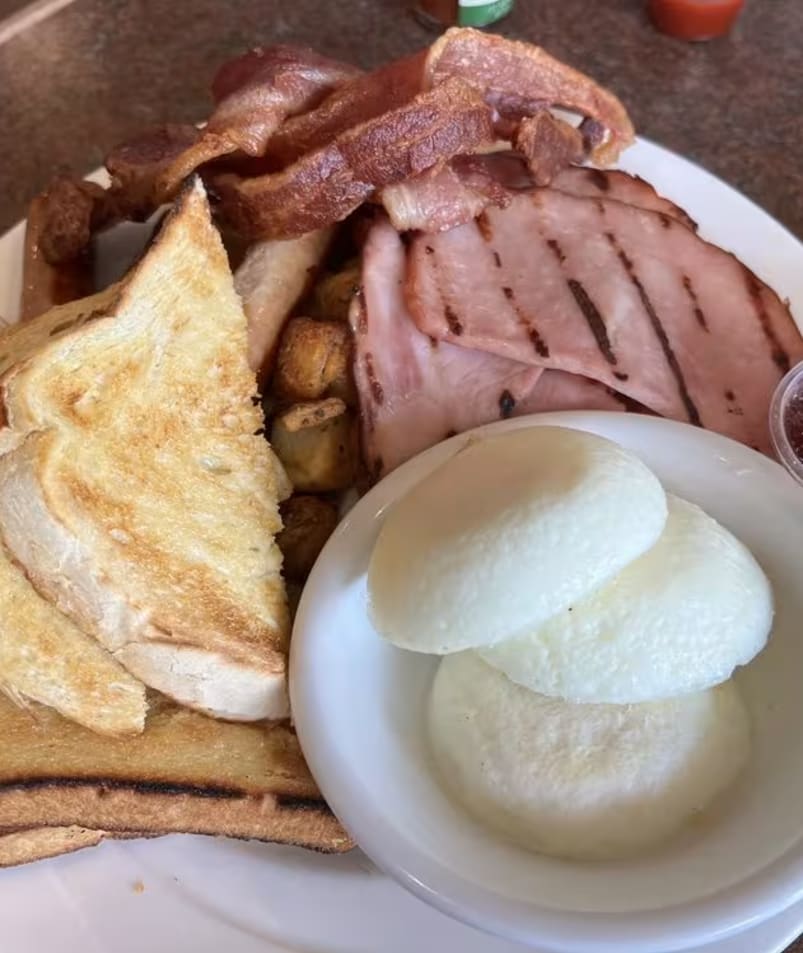 A breakfast plate from The Jammery in Lake Country featuring toast, bacon, ham, poached eggs, hash browns, and small portions of jam.