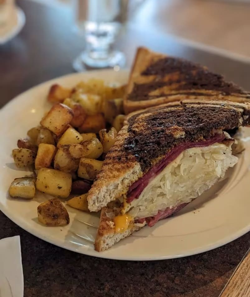 A delicious sandwich with layers of meat and sauerkraut served with a side of roasted potatoes on a white plate at The Jammery in Lake Country, photographed by The Jammery.