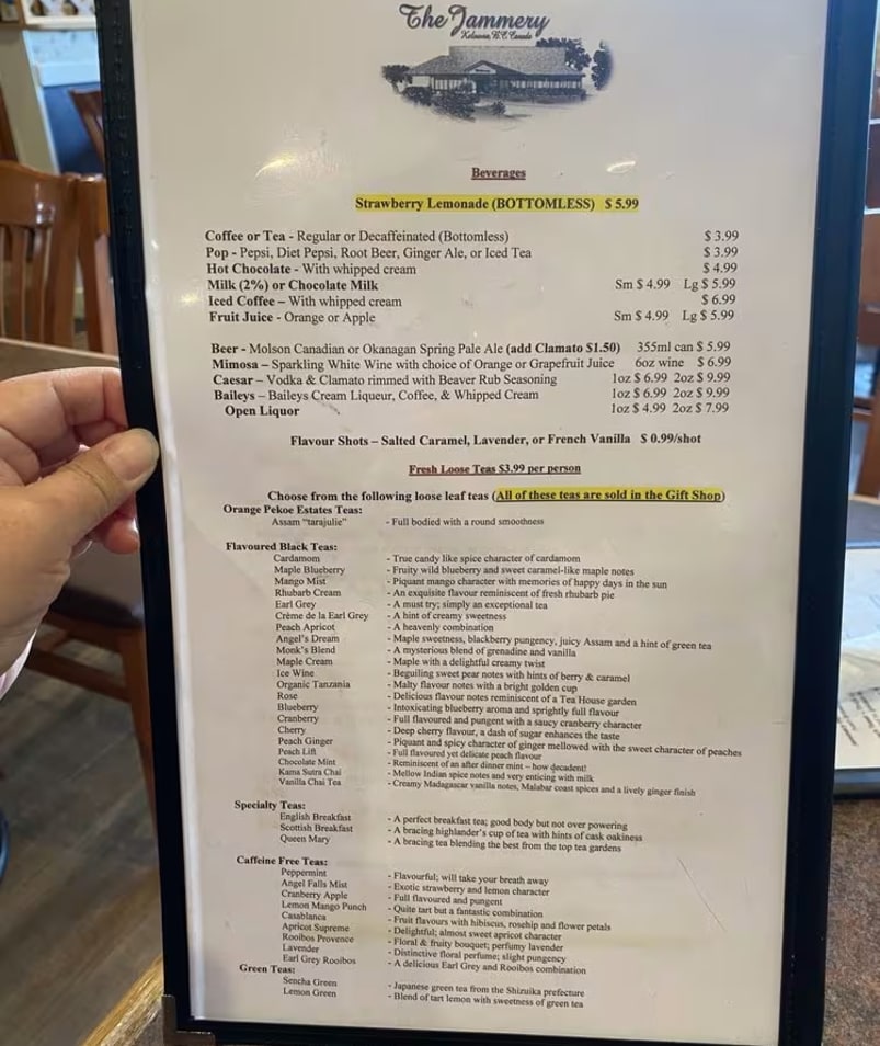 A hand holding a menu from The Jammery in Lake Country, listing various beverages including bottomless strawberry lemonade, coffee, tea, hot chocolate, milk, juice, beer, mimosa, Caesar, Baileys, liquor, flavour shots, and a selection of loose leaf teas.