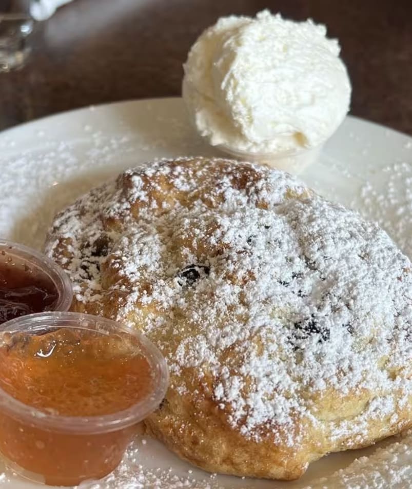 A scone dusted with powdered sugar, served with two small containers of jam and a scoop of whipped cream on a white plate at The Jammery in Lake Country.