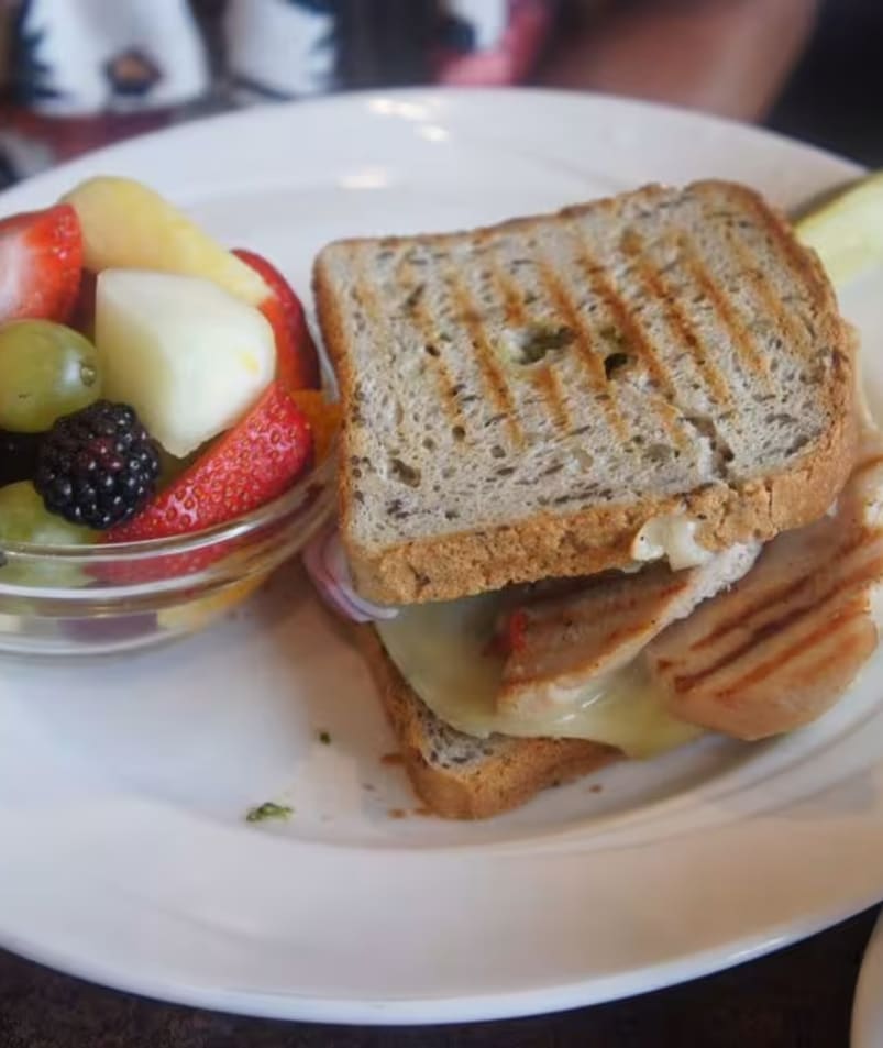 A grilled sandwich on a white plate with a side bowl of mixed fresh fruits at The Jammery in Lake Country.