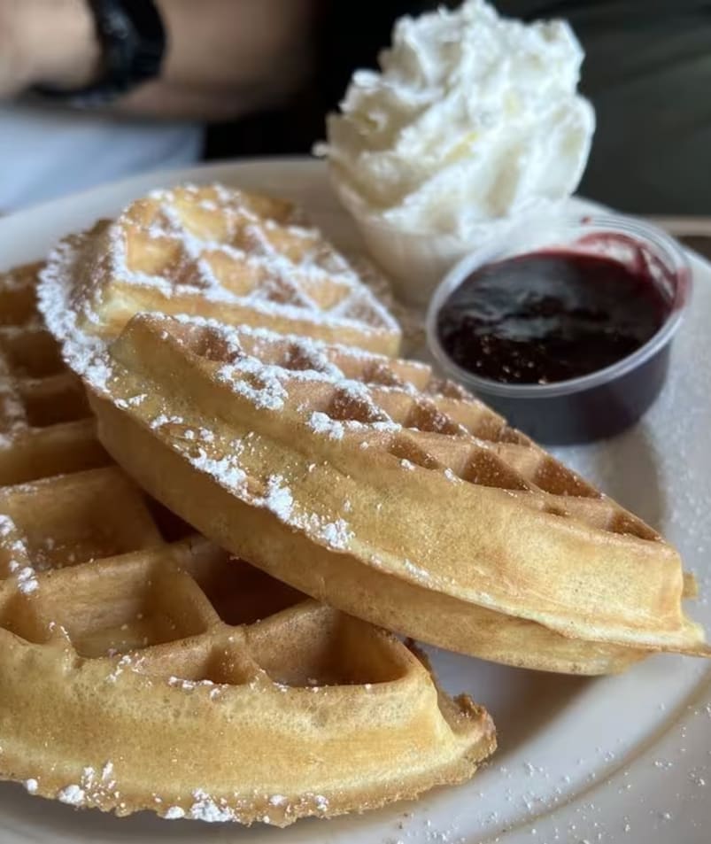 A serving of waffles dusted with powdered sugar, accompanied by a small cup of blackberry sauce and a dollop of whipped cream at The Jammery in Lake Country.