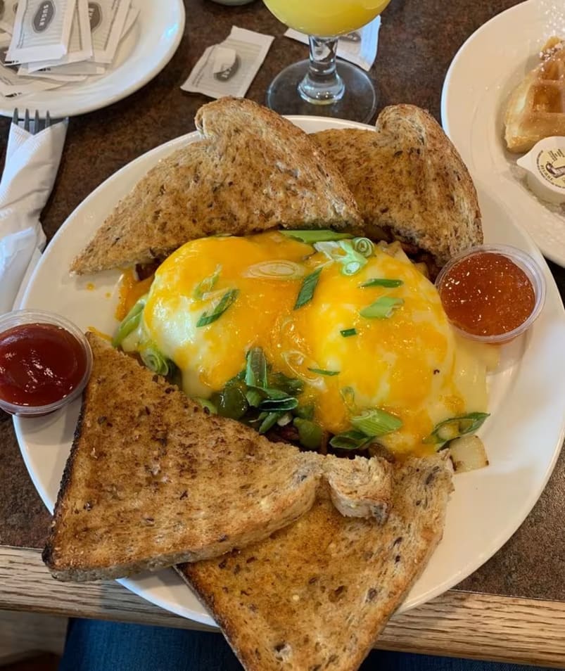A breakfast dish from The Jammery in Lake Country featuring an omelette topped with melted cheese and green onions, with slices of toast, ketchup, and jam, accompanied by a glass of orange juice.