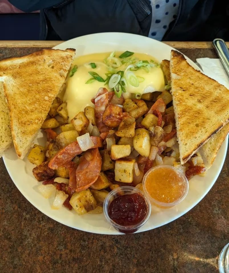 A plate of breakfast food from The Jammery in Lake Country, featuring toast, potatoes, sausage, poached eggs with a creamy sauce, green onions, and small containers of jam and sauce.