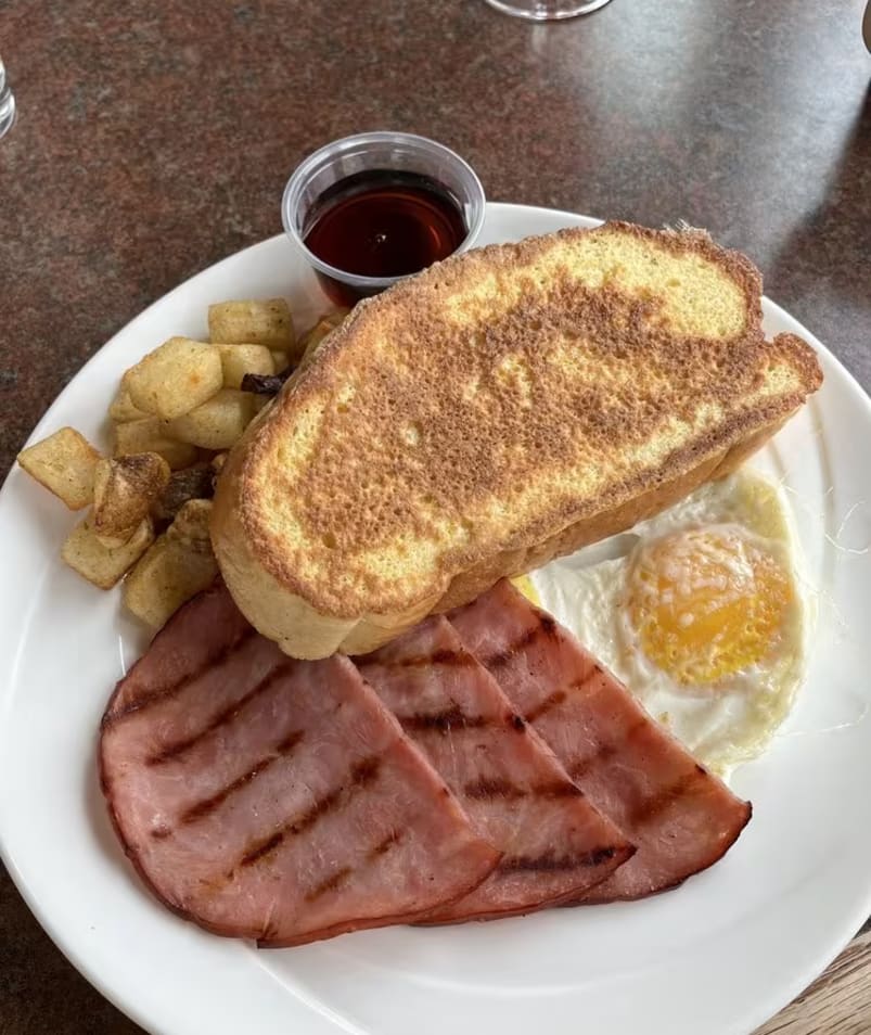A plate of breakfast food from The Jammery in Lake Country, featuring toast, fried eggs, ham, potatoes, and a small cup of syrup.