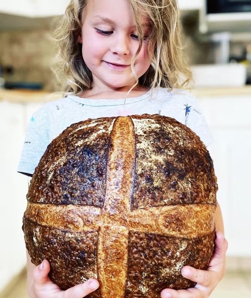 a little girl holding a big loaf of bread in her hands and smiling at the camera while standing in a kitchen