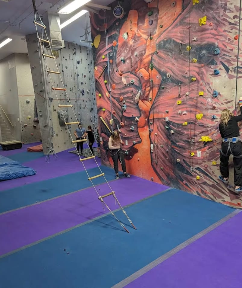 A group of people climbing up the side of a climbing wall in a climbing gym with climbing equipment on the walls.