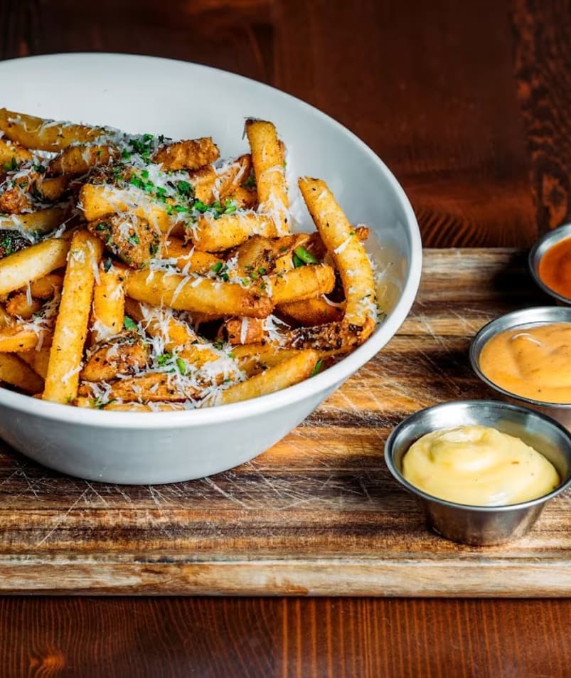 Asymmetrical white bowl with crispy truffle parmesan fries, three dipping sauces on the side.