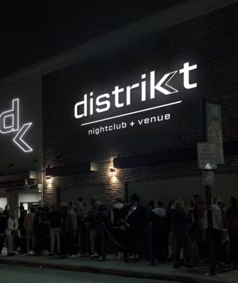 a group of people standing in front of a building at night with the word distrkt lit up on the side of the building.