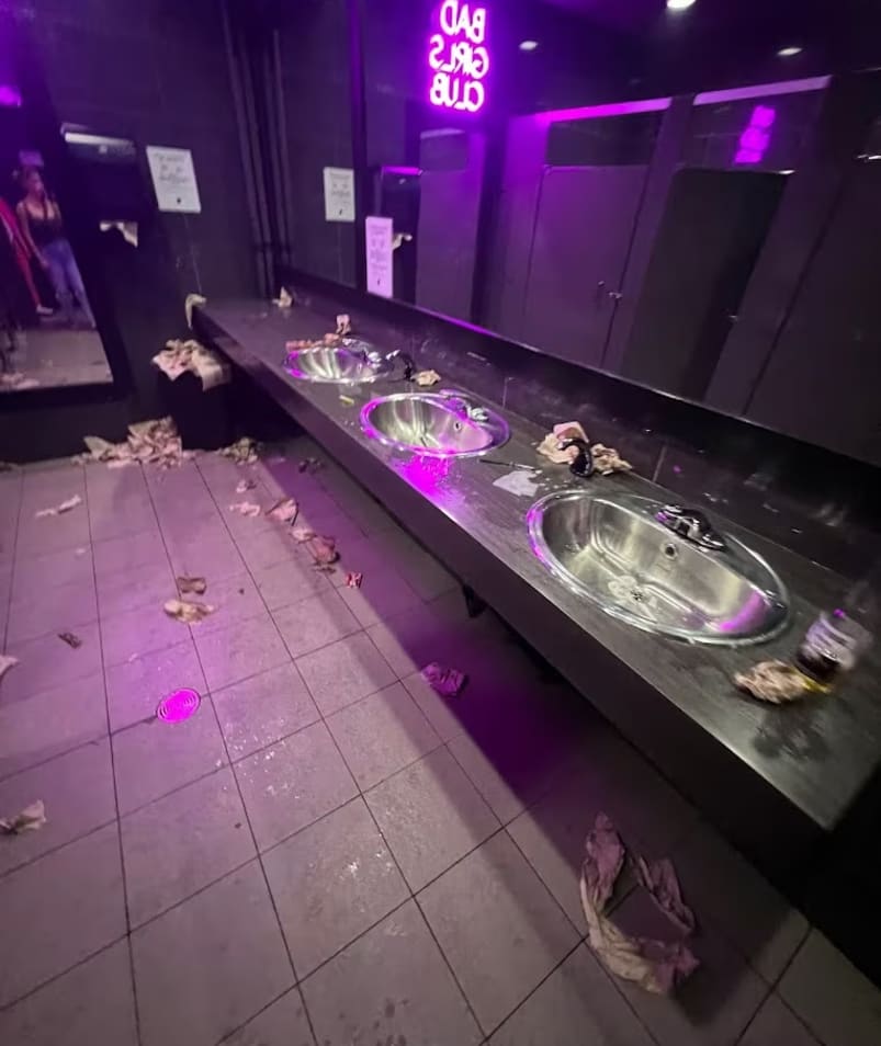 a bathroom with a sink and a lot of trash on the floor and a neon sign above it that says bad girls club