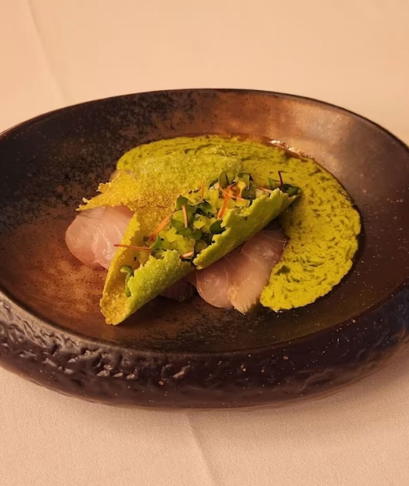 A gourmet dish on a rustic black plate featuring thinly sliced raw fish topped with a vibrant green sauce and garnishes from Erica Jane Restaurant in Kelowna.