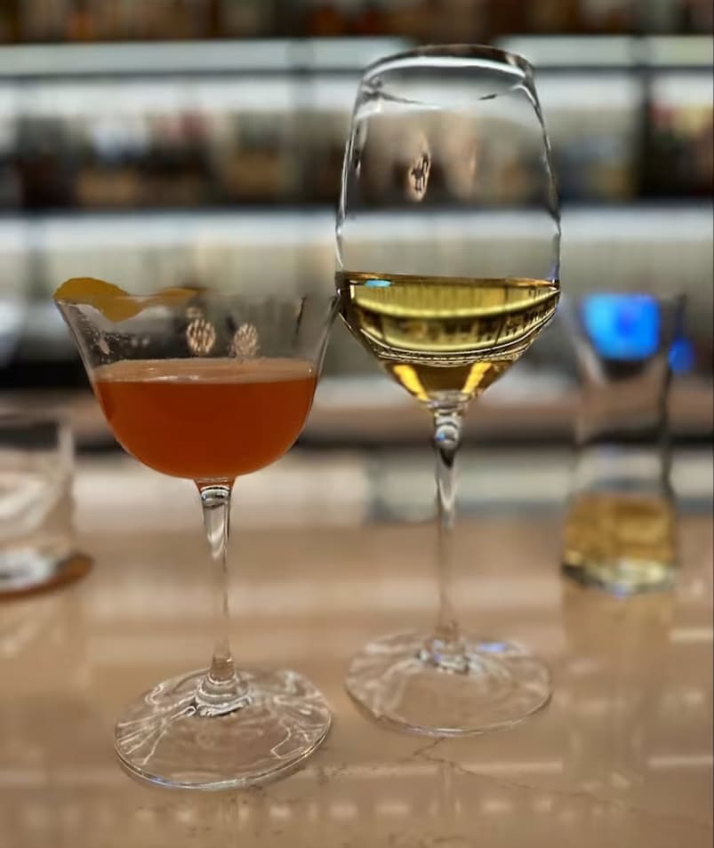 Two elegantly served drinks, one a cocktail with a citrus twist and the other a glass of white wine, on a polished bar counter at Erica Jane Restaurant in Kelowna.