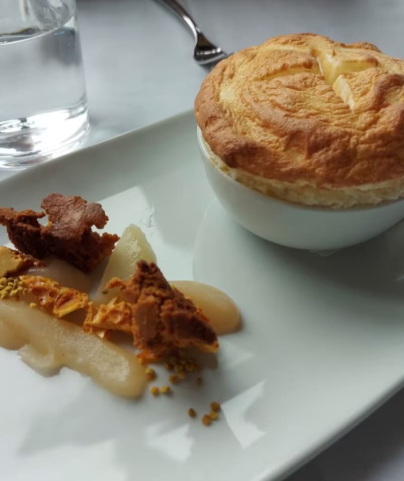 A dish with a puff pastry in a white ramekin, garnished with delicate food items, served on a white plate next to a glass of water on a table at Erica Jane Restaurant in Kelowna.