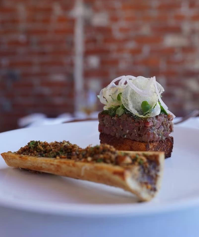 A gourmet dish featuring finely prepared meat garnished with herbs and thinly sliced vegetables, served artfully on a white plate at Erica Jane Restaurant in Kelowna.