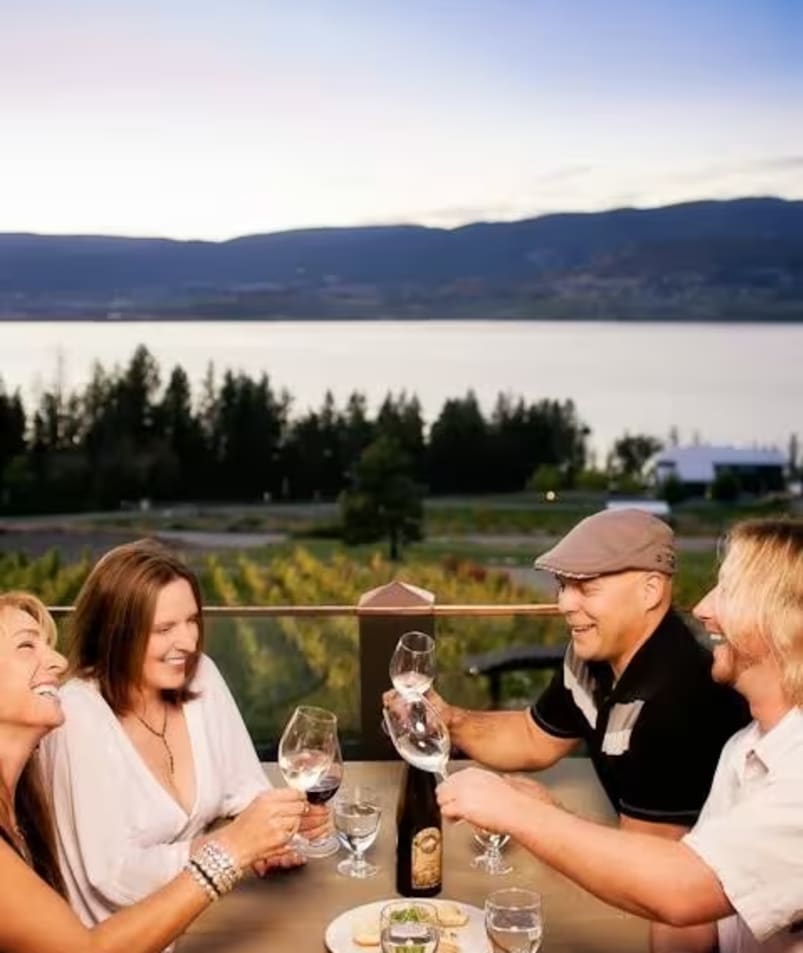 A group of people sitting at a table with wine glasses in their hands and a bottle of wine in the other hand at IMAGINE Organic Bistro, with a view of a lake and mountains in Kelowna.