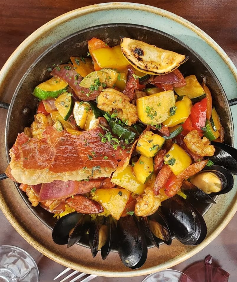 a bowl of food with mussels and other food items on a table with wine glasses and utensils