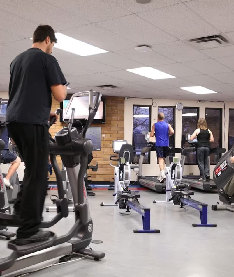 A group of people in a gym with treadmills and exercise bikes in the foreground and a man on a cell phone in the middle of the room.