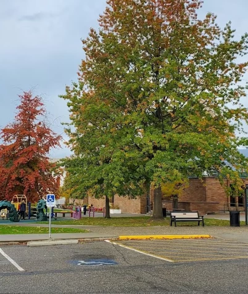 a parking lot with a tree in the middle of it and a playground in the back of the lot at Parkinson Recreation Centre in Kelowna