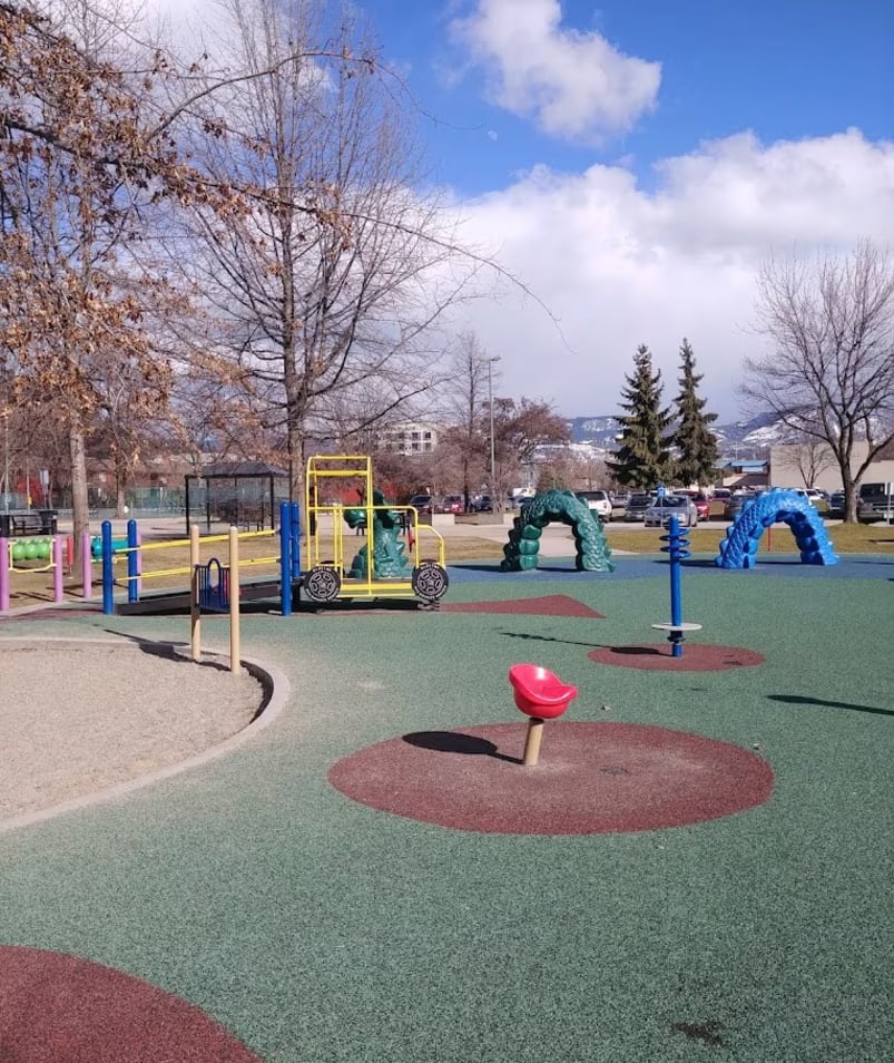 A park with a playground and a play area with swings, slides, and play structures under a blue sky at Parkinson Recreation Centre in Kelowna.