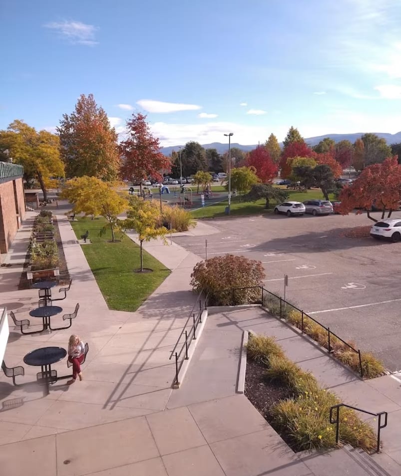 A high point of view showing the Parkinson Recreation Centre parking lot in Kelowna with trees, playground, and mountains in the background.