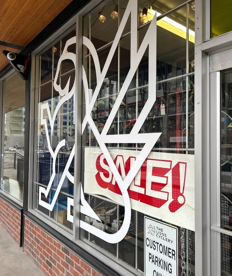 A storefront with a sign that says SALE! in the front window and The Lions Cyclery CUSTOMER PARKING ONLY on the side window.