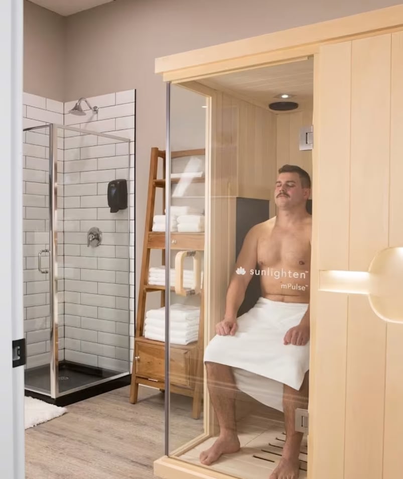 A man sits in a wooden infrared sauna cabin at The Recovery Spa in Kelowna, with a tiled shower and wooden shelf with towels in the background.